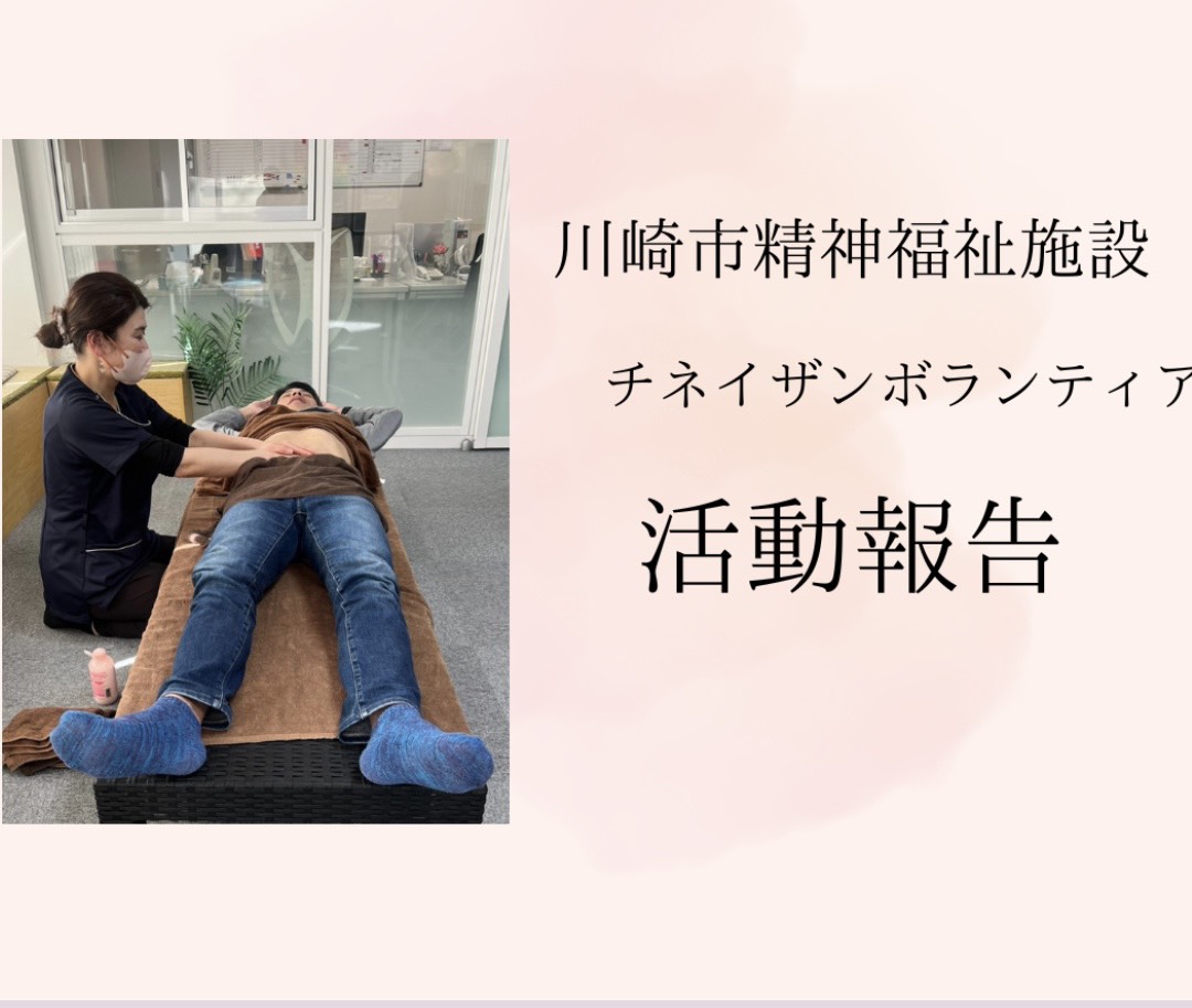 マッサージベッドに横たわる利用者の腹部に、施術者が優しく手を添えてチネイザン（内臓マッサージ）を行っている様子。施術者はマスクと制服を着用し、落ち着いた室内で丁寧にお腹をほぐしている。背景には観葉植物や仕切りがあり、福祉施設でのボランティア活動の一場面を示す写真。