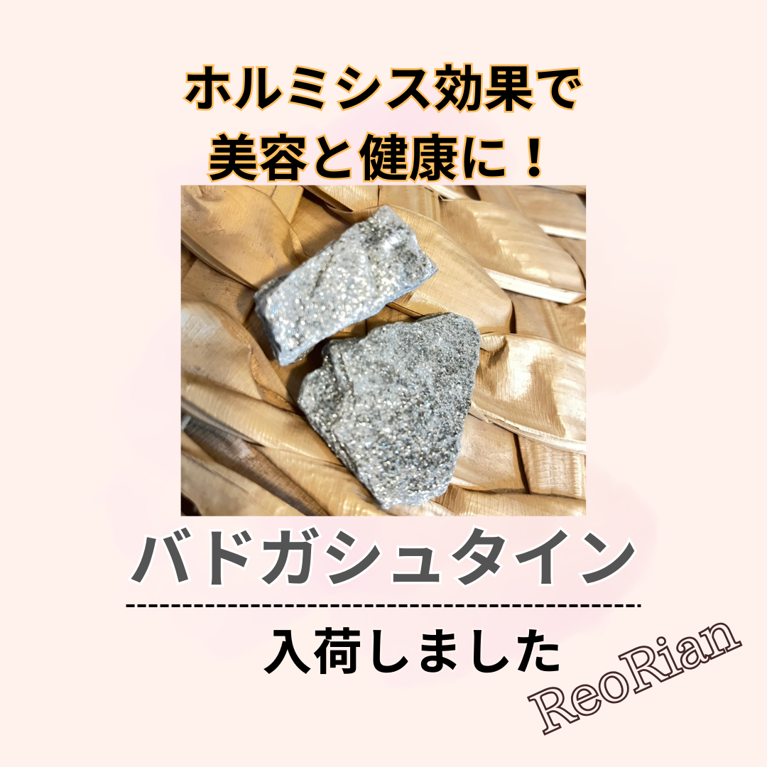 編み目のある台の上に置かれた2つの灰色の鉱石の写真。鉱石は金属光沢のある質感で、上部には『ホルミシス効果で美容と健康に! バドガシュタイン入荷しました ReoRian』というテキストが配置されている。