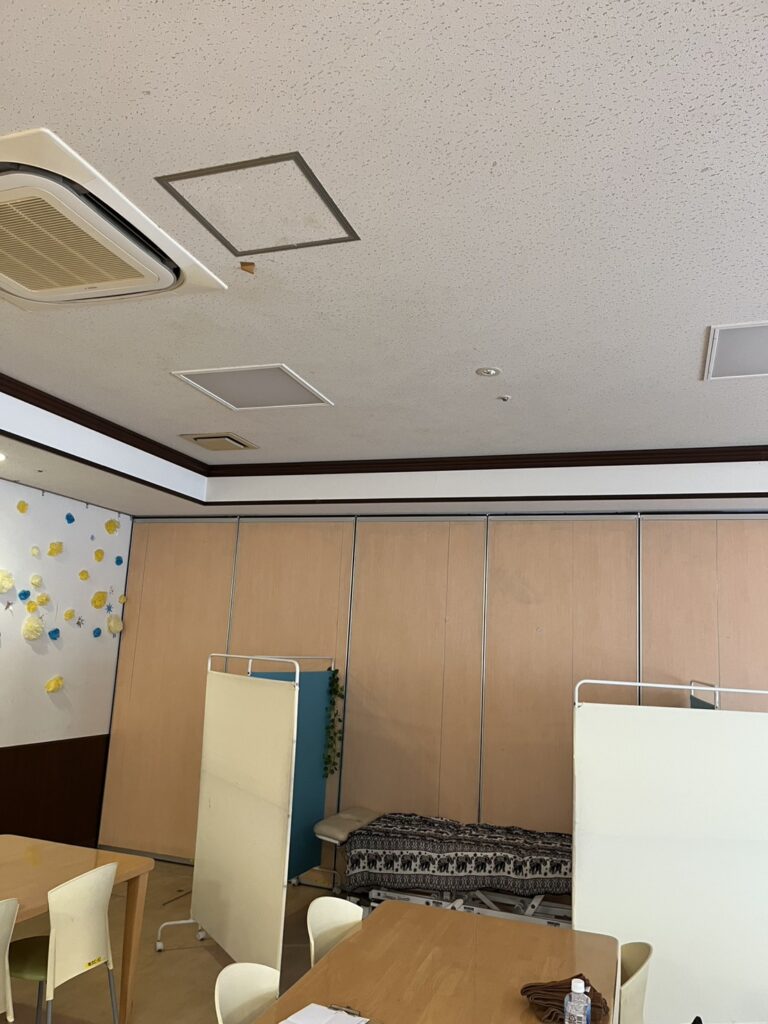「室内の多目的スペース。白い天井に天井埋込型エアコンと照明パネルがあり、壁は明るい木目調。折りたたみ式のパーテーション、テーブルと椅子、柄付きのカバーがかかったベンチまたは施術用ベッドが配置されている。左側の壁には黄色と青の紙製の花飾りがある。」