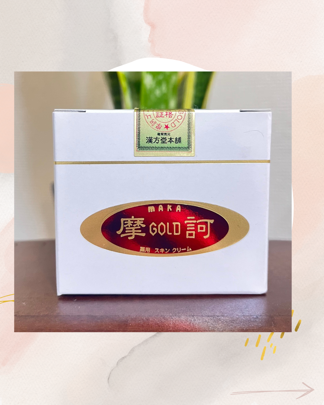 「白い箱型のパッケージが木のテーブルに置かれており、中央に赤と金の楕円形ロゴ『MAKA GOLD』と日本語の文字が印刷されている。上部には金色のラベルがあり、背景にはぼかした緑の植物が写っているスキンケア商品の写真。」