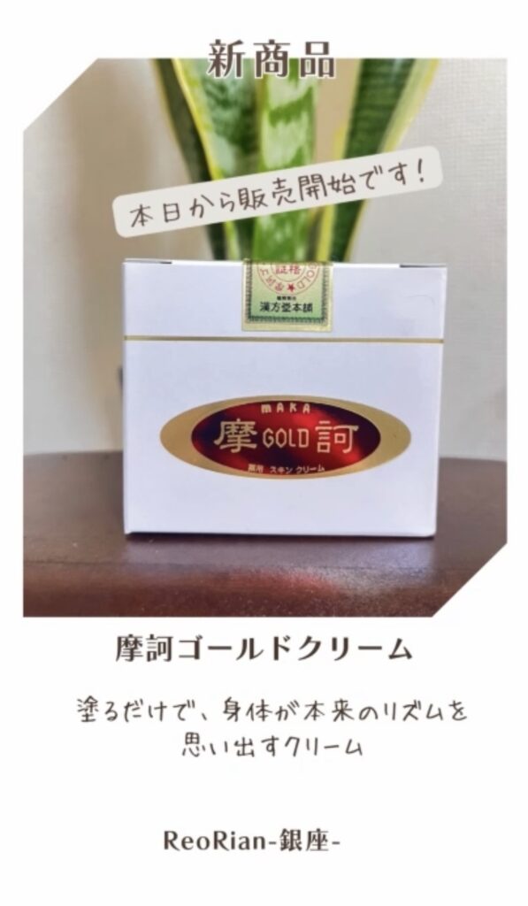 木のテーブルの上に、摩訶ゴールドクリームの白い箱が置かれている。箱には赤と金のラベルがあり、背景には緑の植物が写っている。上部に『新商品 本日から販売開始です！』という文字が表示されている。」